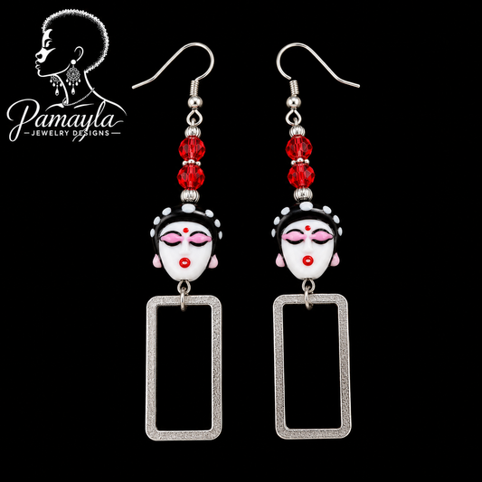 Pair of earrings with face designs and red beads on a black background, featuring the brand 'Pamayla Jewelry Designs'.